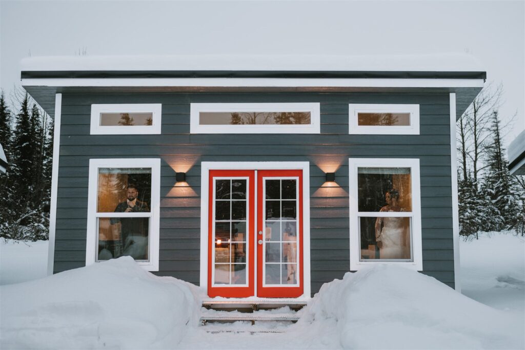 A snow-covered modern cabin with a dark exterior and red-framed doors offers an ideal setting for a winter elopement. Tall windows flank the entrance, allowing glimpses of the trees in the background.