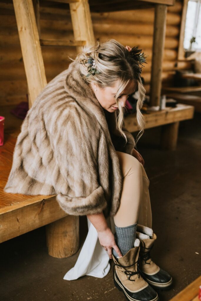 A woman in a fur coat and flower crown sits on a wooden bench, preparing for a winter elopement by putting on her boots in a rustic setting.