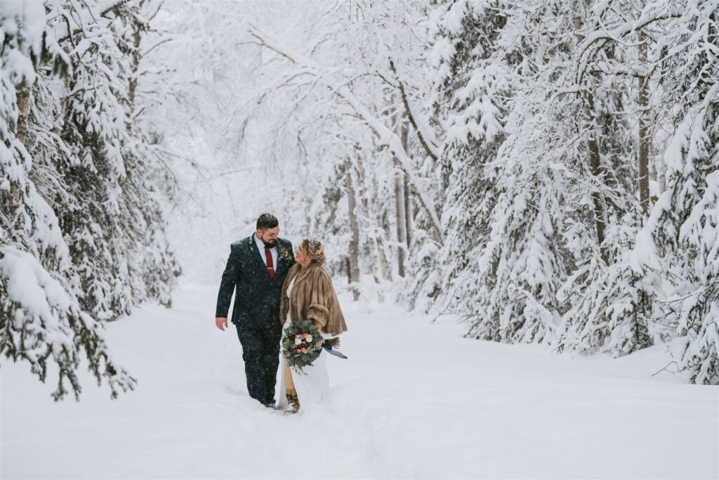 A couple in formal attire walks through a snowy forest, surrounded by snow-covered trees. The woman carries a bouquet, capturing the magic of a winter elopement.