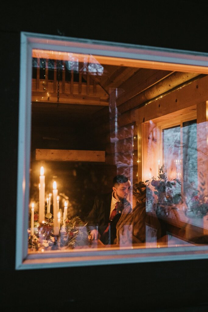 Two people are sitting inside a dimly lit room, viewed through a frosty window. Candles flicker on the table, casting a cozy glow around them, setting the perfect scene for an intimate winter elopement.