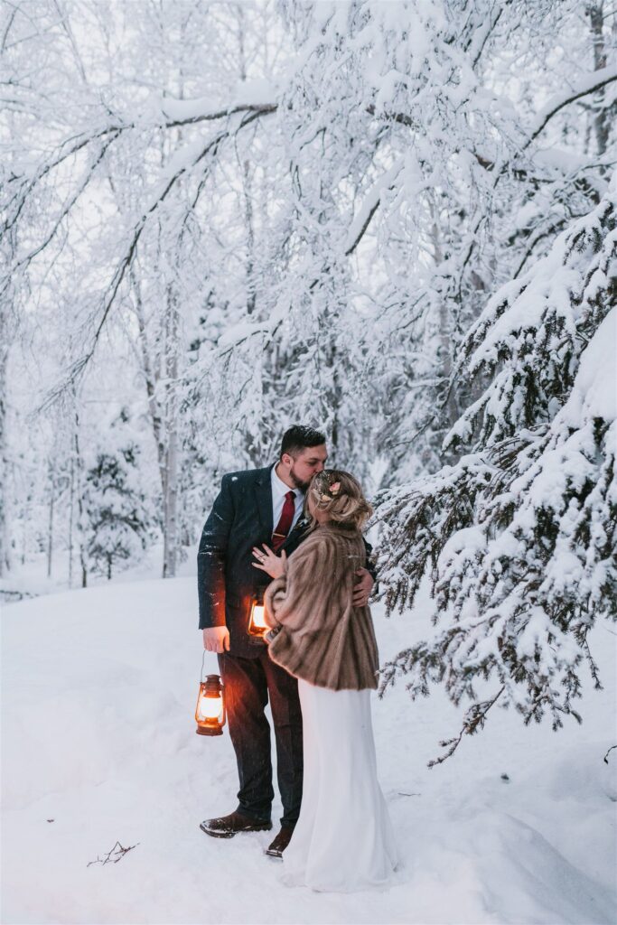 In a snowy forest, a couple embraces, lanterns glowing softly in their hands. The scene, perfect for a winter elopement, is framed by snow-laden trees and an ethereal quiet that envelops them.