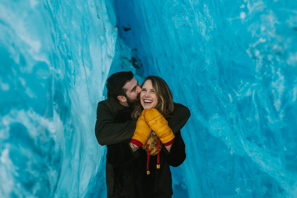 A couple in winter clothing stands inside a blue ice cave, wrapped in the magic of a winter elopement. The man embraces the smiling woman.