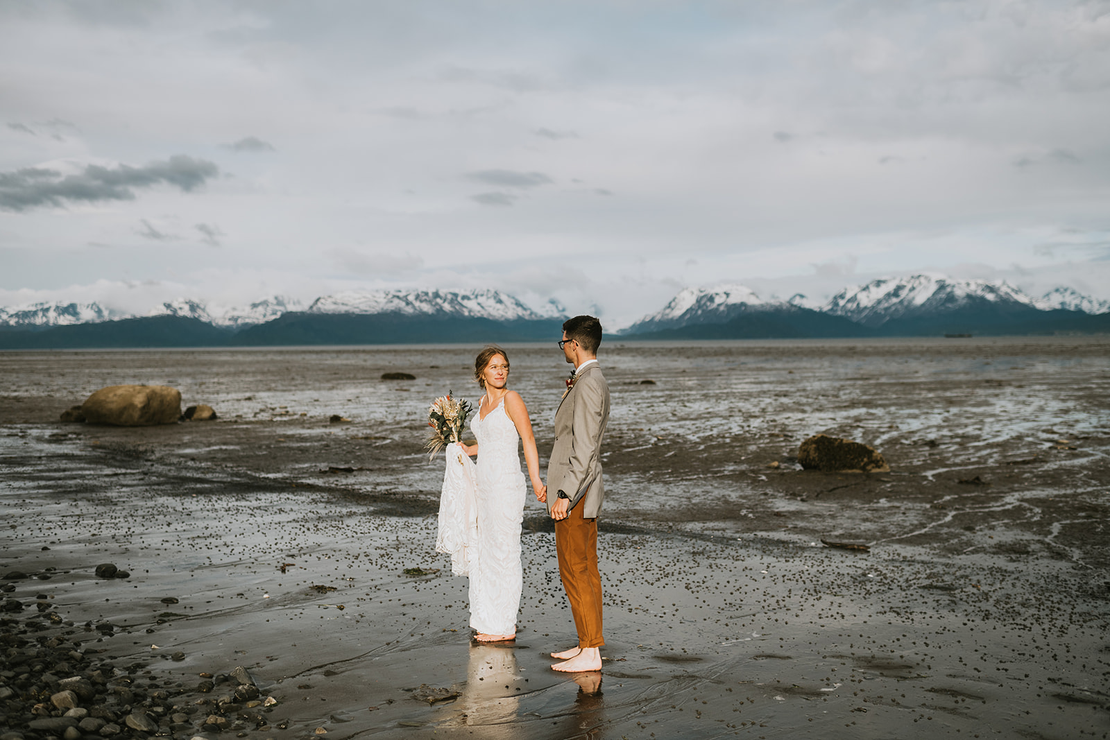 In one of the picturesque Alaska coastal towns, a bride in a white dress and groom in a suit stand barefoot on a sandy beach with majestic mountains towering in the background.