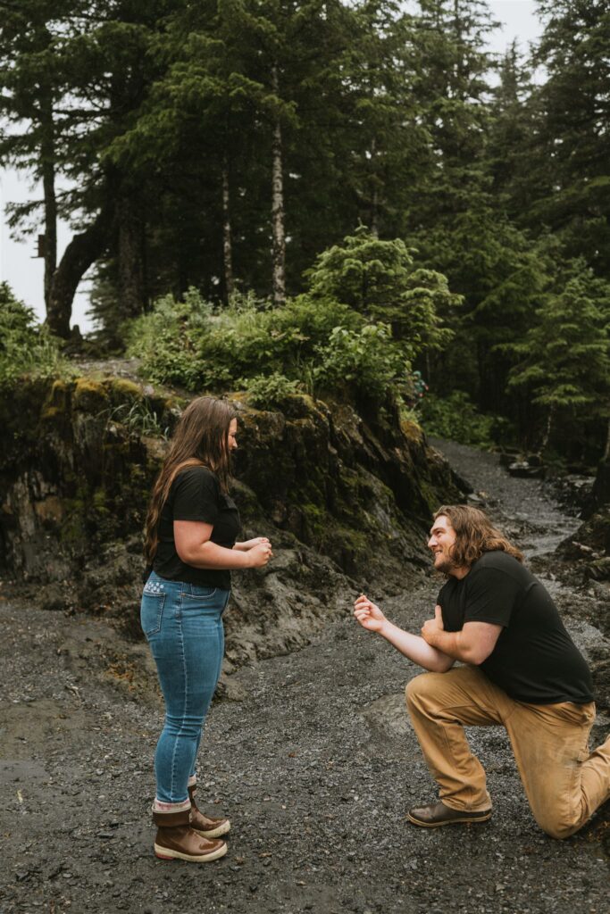 A man kneels on one knee proposing to a woman on a gravel path in a forested area, offering one of the most romantic proposal ideas surrounded by nature.