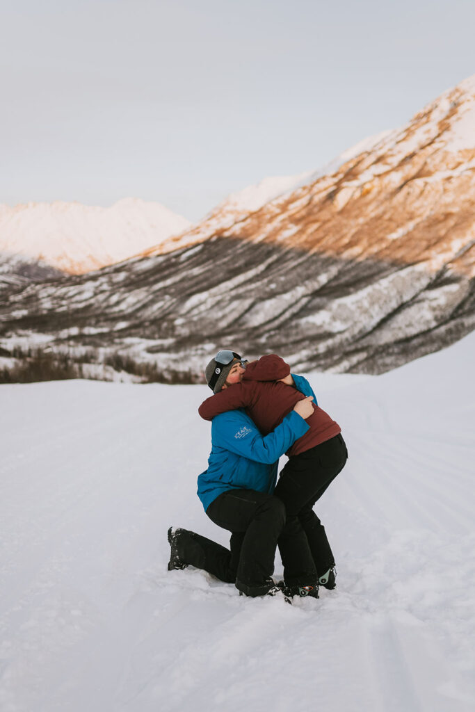 Two people in winter jackets embrace each other on a snowy mountain landscape, with snow-covered peaks and gentle sunlight in the background—an unforgettable scene for romantic proposal ideas.