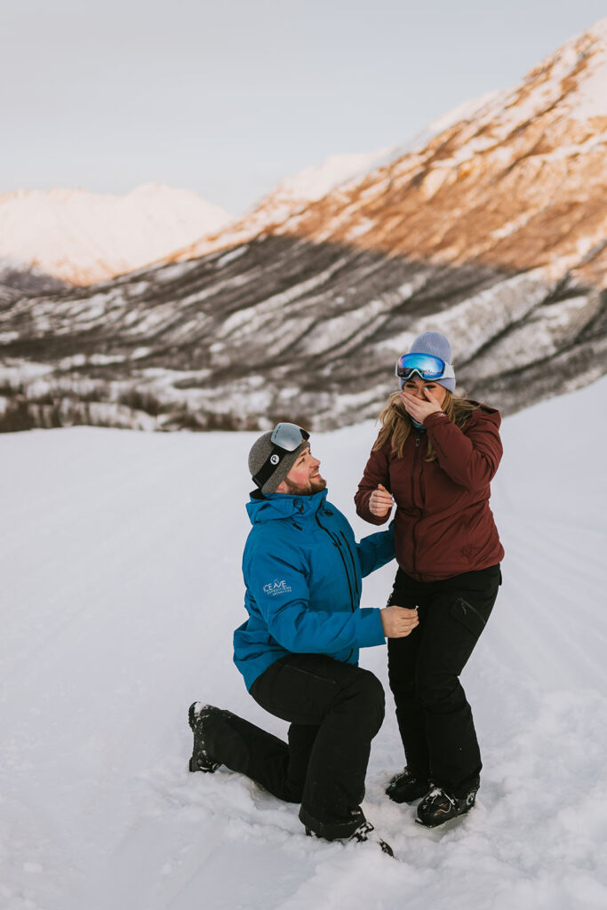 A person kneels in the snow proposing to another, both dressed in winter clothing with snowy mountains in the background—one of the most romantic winter proposal ideas.