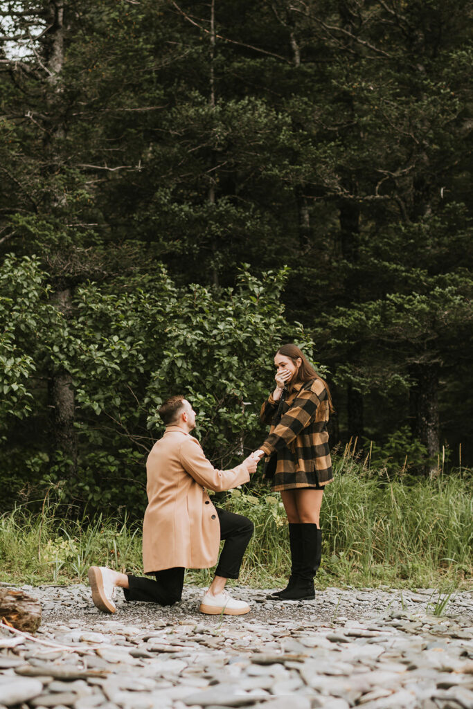 A man kneels and proposes to a surprised woman outdoors on rocky ground, with trees and greenery in the background—capturing one of the most romantic proposal ideas in nature.