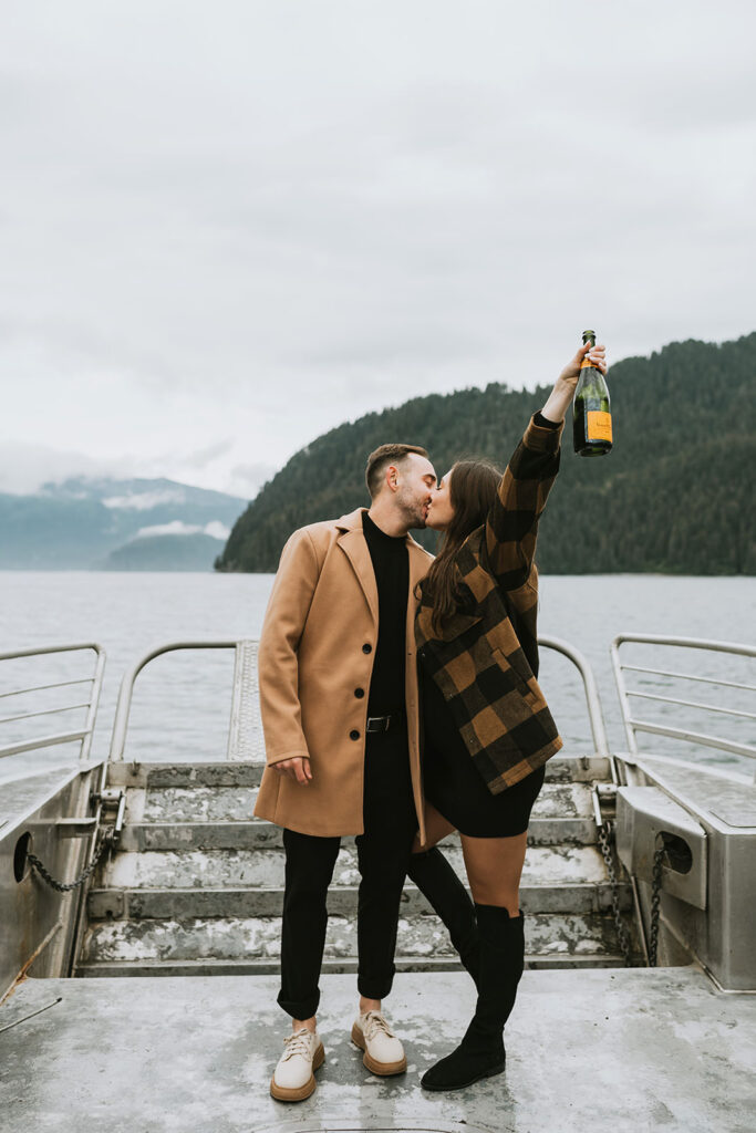 A couple stands on a boat dock, kissing, while the woman holds up a bottle—one of those dreamy proposal ideas with mountains and water in the background under a cloudy sky.