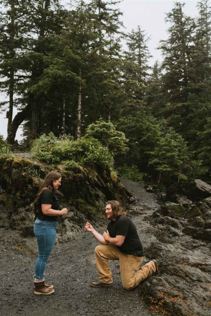A man kneels and proposes to a woman on a forested, rocky trail—one of the most romantic proposal ideas; both wear casual clothes, surrounded by towering trees.