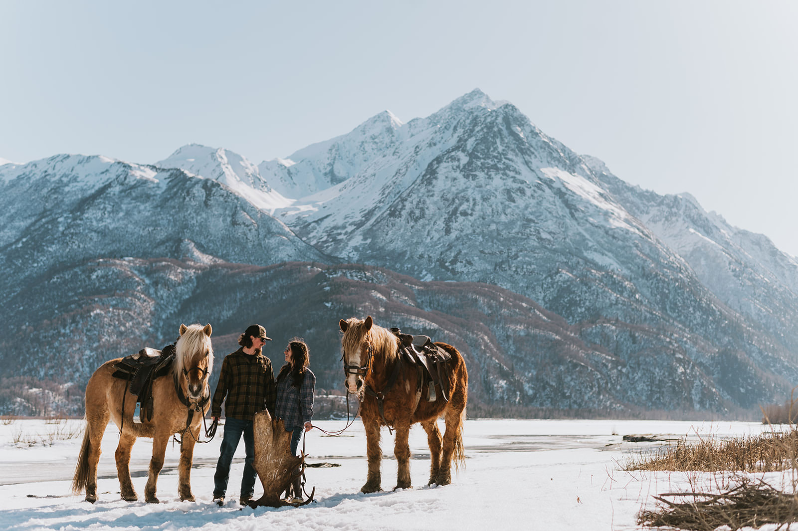 A man and woman stand with two saddled horses on a snowy field, tall mountains in the background under a clear sky—an unforgettable setting for romantic proposal ideas.