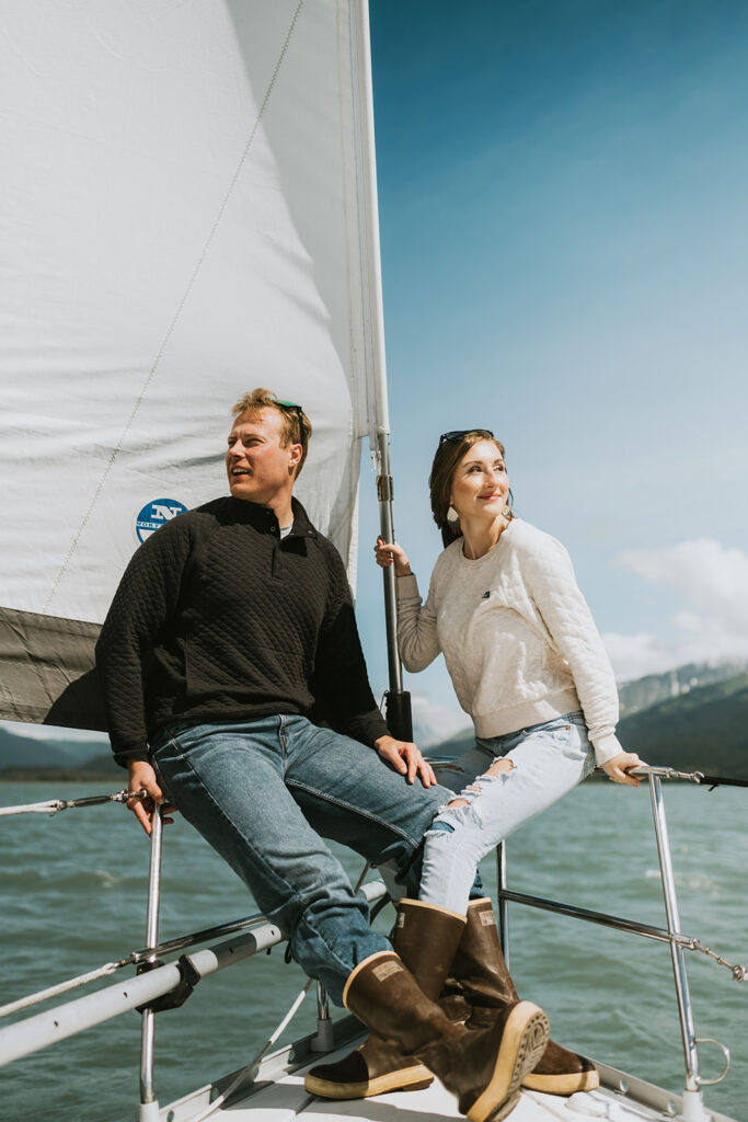 A man and woman sit at the bow of a sailboat on the water, both looking off into the distance—an idyllic setting that inspires romantic proposal ideas. The sky is clear, with mountains visible in the background.