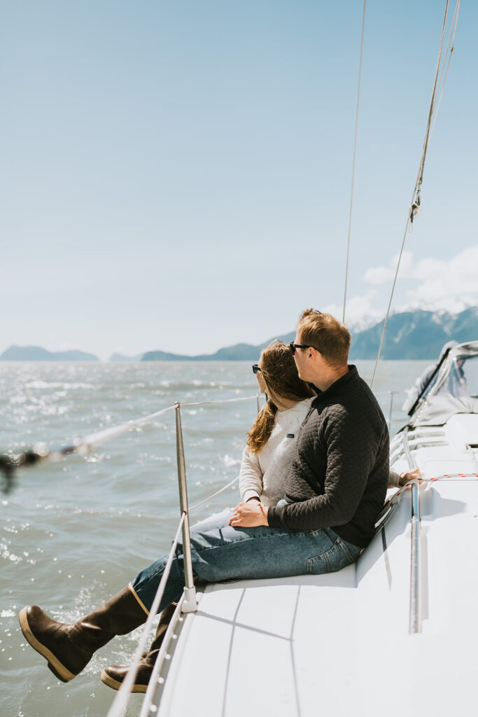 A man and woman sit together on the edge of a sailboat, looking out at the water and distant mountains under a clear sky—one of the most romantic proposal ideas.