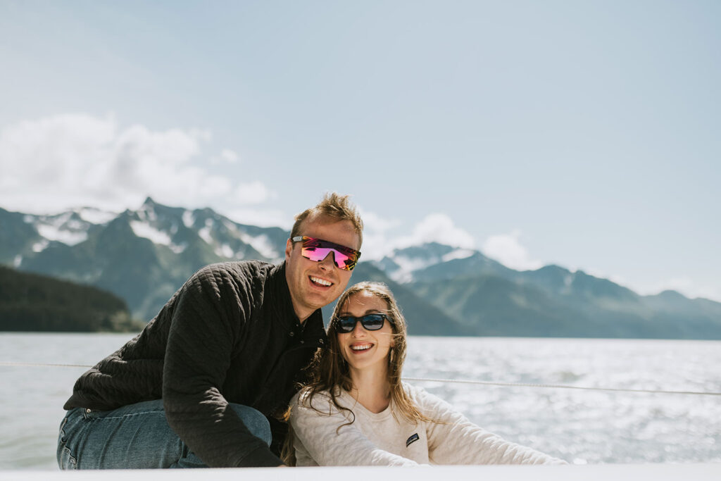 A man and woman wearing sunglasses smile while sitting on a boat with mountains and water in the background, making it a perfect setting for romantic proposal ideas.