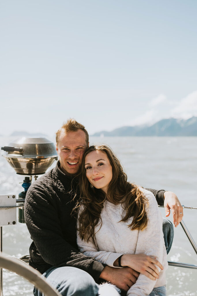 A man and woman sit closely together on a boat, smiling at the camera with the ocean and mountains in the background—perfect inspiration for romantic proposal ideas.