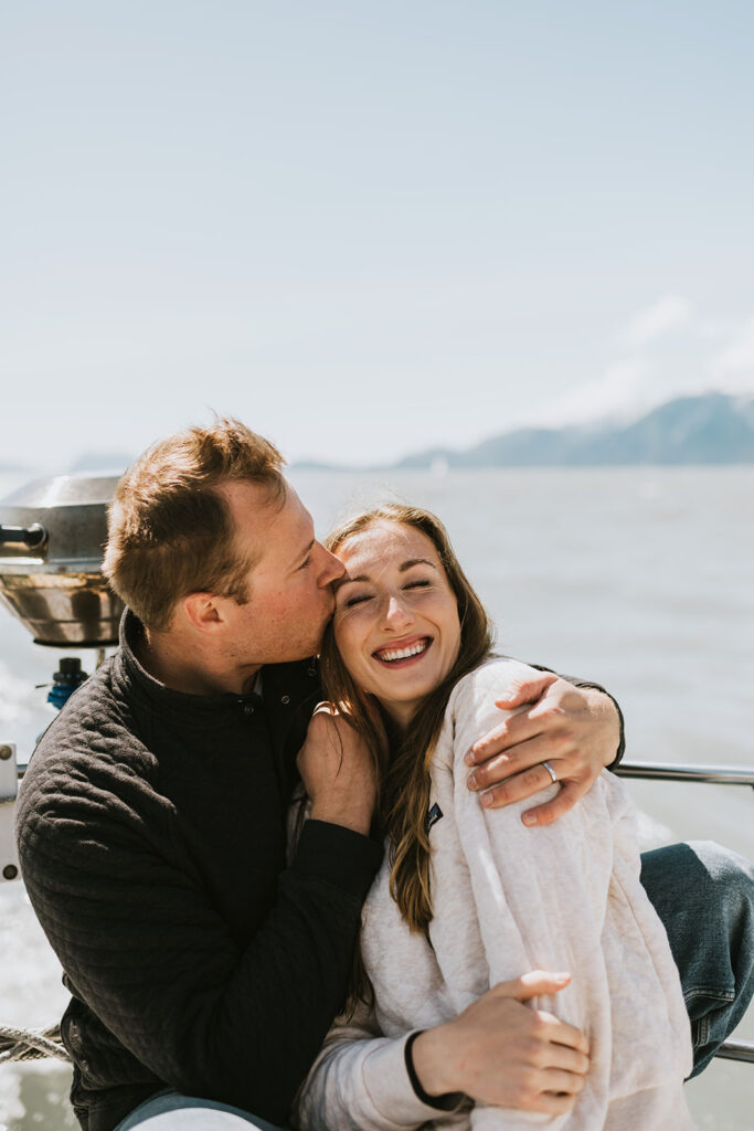 A man kisses a smiling woman on the cheek as they sit together on a boat, with water and mountains in the background—creating one of those unforgettable proposal ideas.