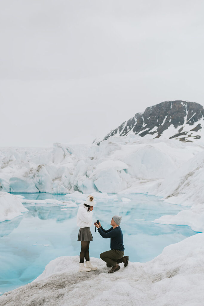 A person kneels to propose beside a blue glacial lake surrounded by snow and ice, with a rocky mountain in the background—one of the most breathtaking proposal ideas.