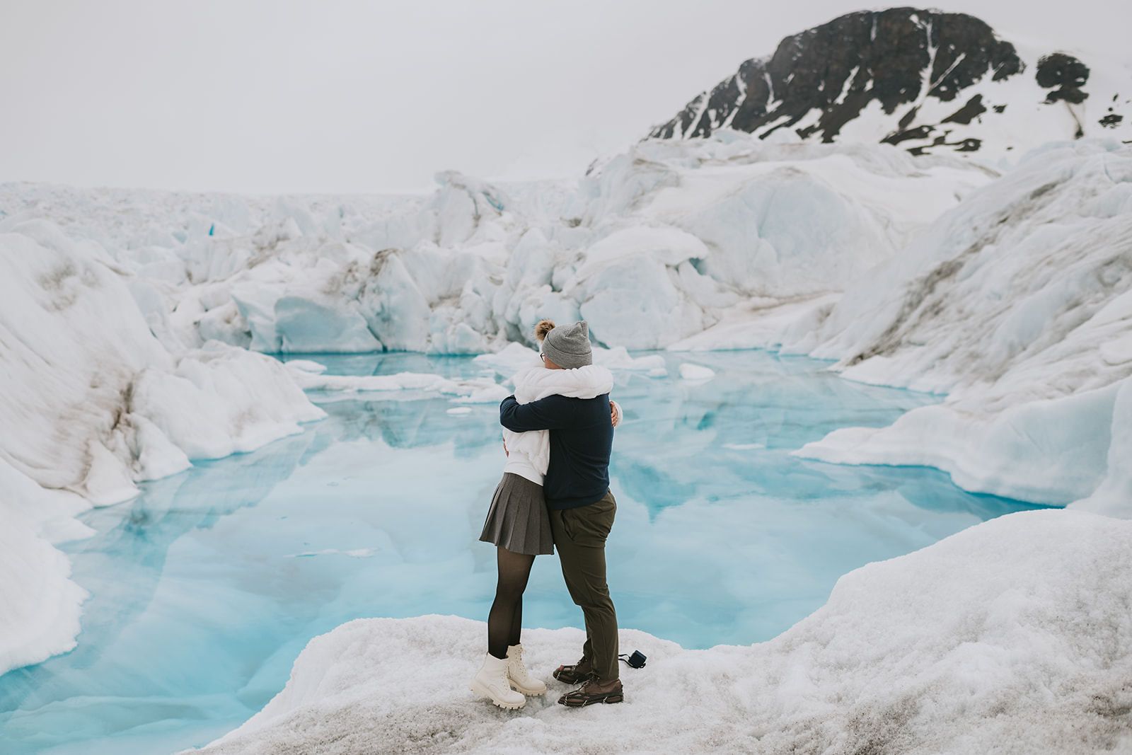 Two people embrace on snowy ground beside a bright blue glacial pool, surrounded by ice formations and a rocky mountain—an unforgettable setting for romantic proposal ideas.