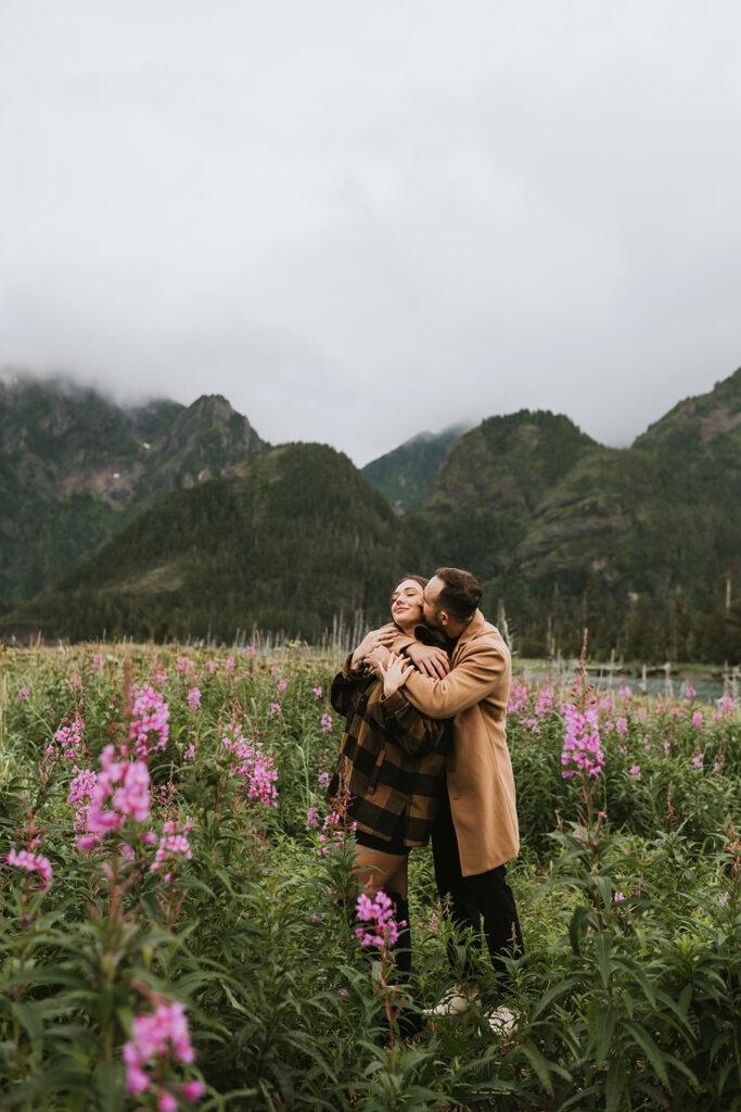 A couple stands in a field of pink flowers with mountains and cloudy skies behind them. The man kisses the woman's cheek as she smiles, capturing one of the sweetest proposal ideas for nature-loving romantics.