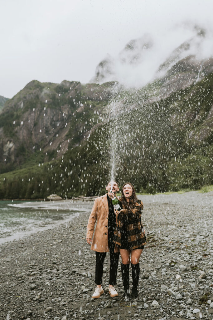 Two people standing on a rocky beach pop a champagne bottle, the spray captured mid-air—a magical moment for unforgettable proposal ideas, with mountains and water stunningly visible in the background.