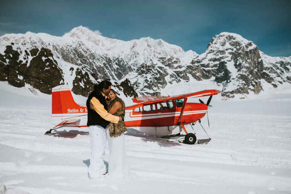 A couple stands embracing on a snowy landscape in front of a red and white small airplane, with snow-covered mountains in the background—one of the most breathtaking proposal ideas.