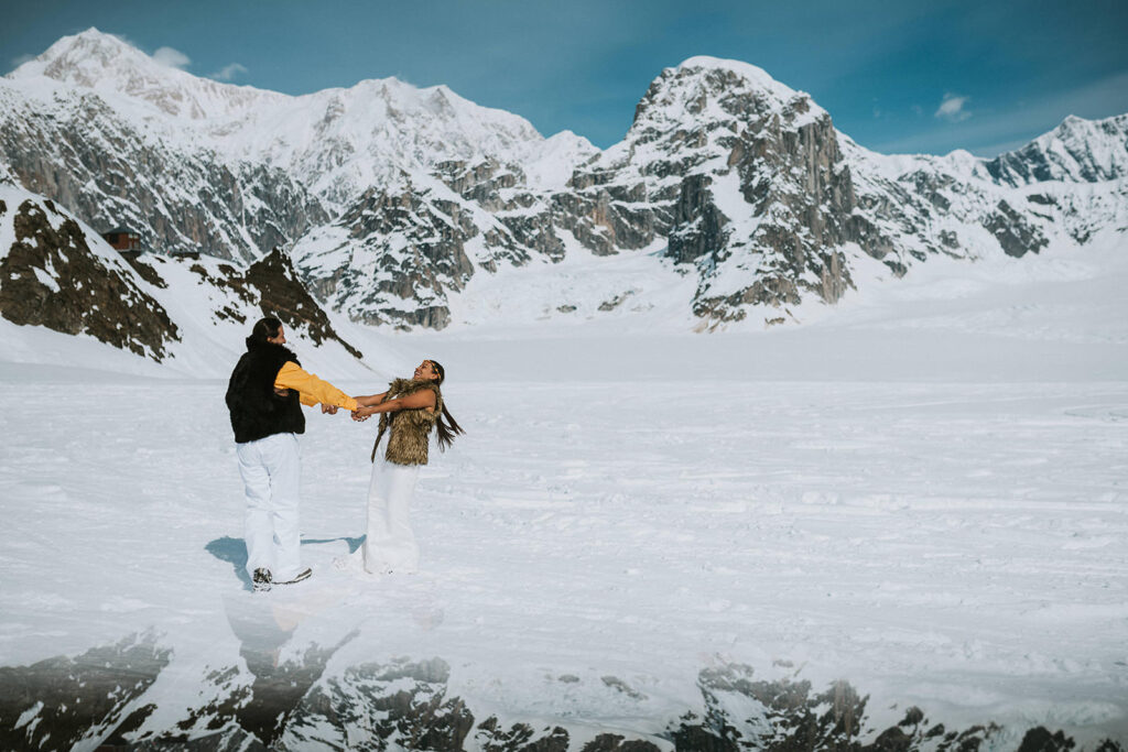 Two people hold hands and spin on a snow-covered landscape, surrounded by large, rugged mountains under a clear blue sky—an unforgettable spot for unique proposal ideas.