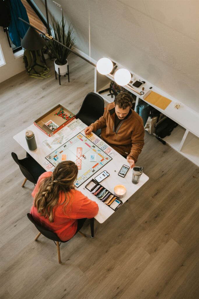 Two people sit at a table playing Monopoly in a modern, well-lit room. Snacks, drinks, and a smartphone are on the table. Captured from above, the scene holds an unexpected lakeside elopement vibe through its relaxed, intimate atmosphere.