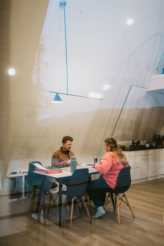 Two people sit at a dining table playing Monopoly in a modern kitchen, viewed through a window with light reflections—capturing the playful spirit of a lakeside elopement at home.
