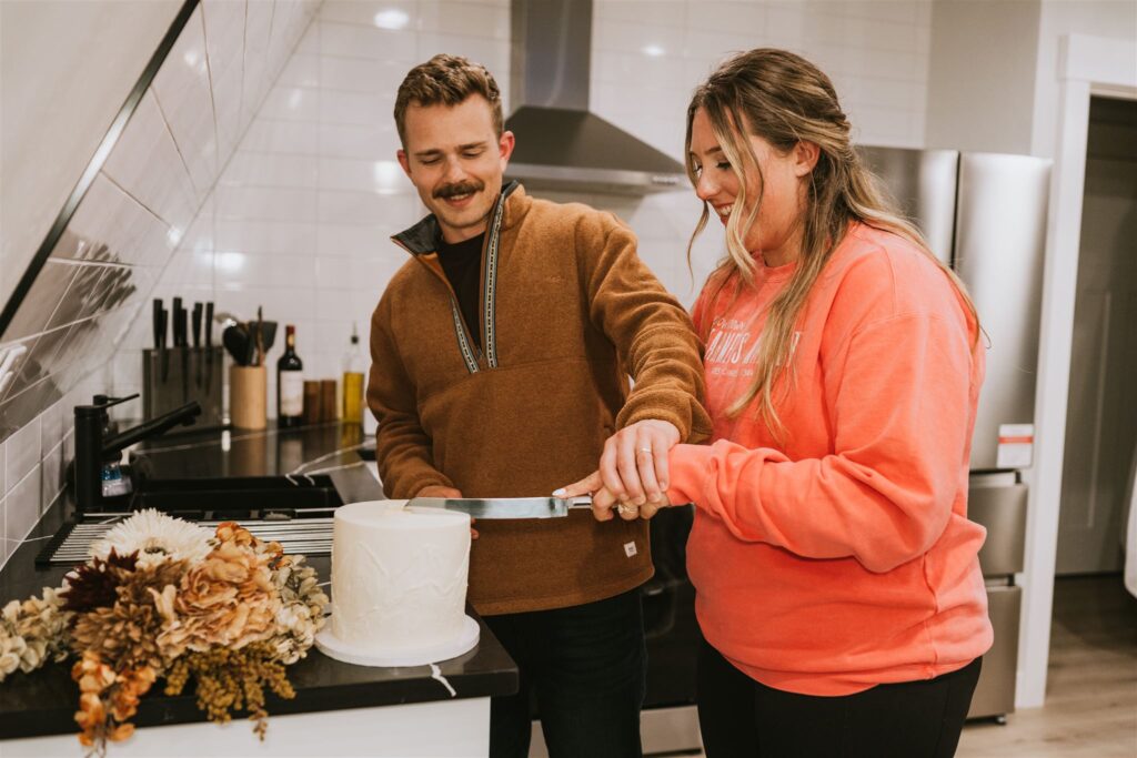 Two people stand in a kitchen, slicing a white cake together after their lakeside elopement; dried flowers are arranged beside the cake on the counter.