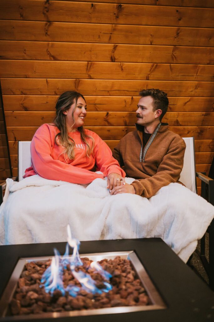 A woman and man sit together on a chair outdoors during their lakeside elopement, wrapped in a white blanket, holding hands and smiling, with a fire pit burning in the foreground.