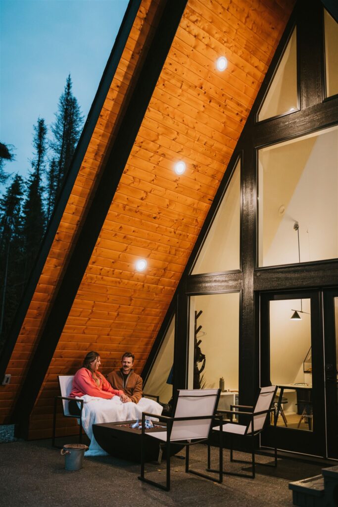 Two people sit together on a patio outside an A-frame cabin at dusk, wrapped in a blanket, outdoor lights illuminating the wooden exterior—the perfect cozy setting for a lakeside elopement.