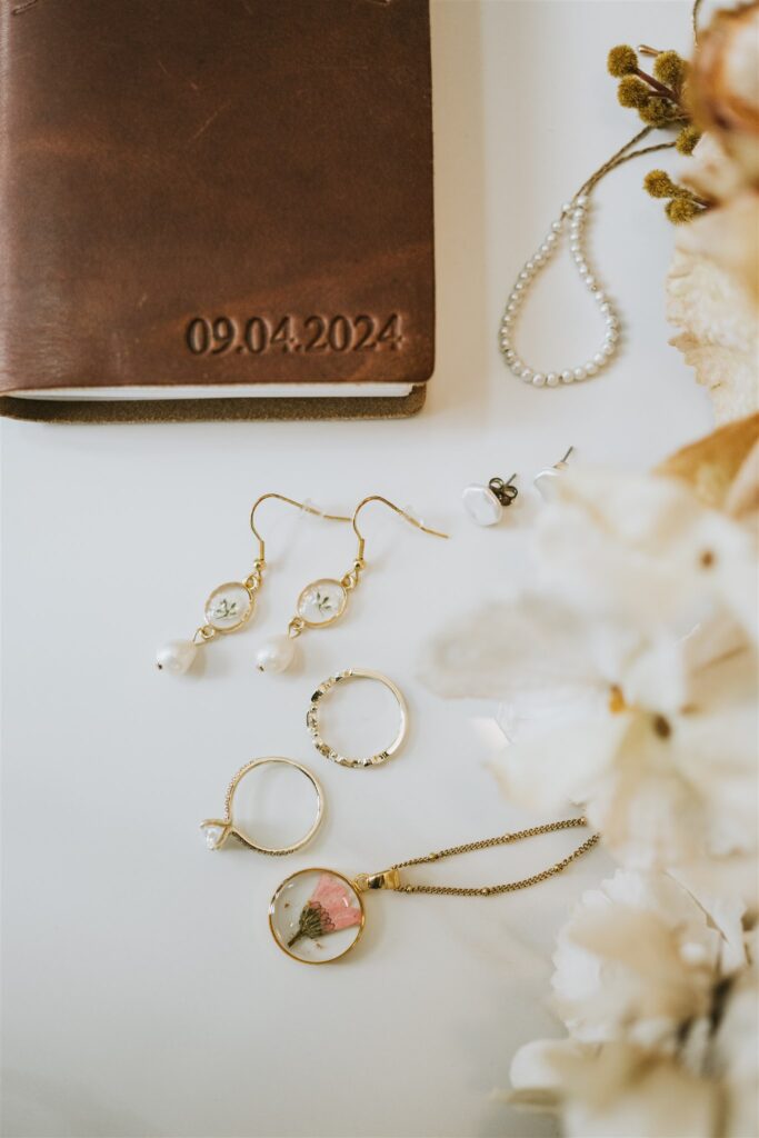Assorted jewelry, including earrings, rings, a bracelet, and a pendant necklace—perfect accents for a lakeside elopement—displayed next to a brown leather-bound planner embossed with the date 09.04.2024.