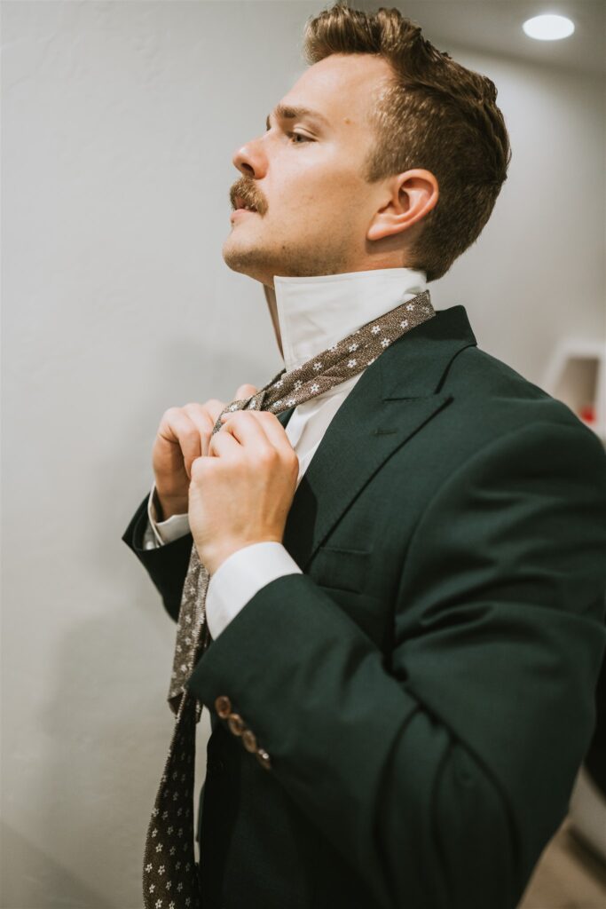 Man in a green suit tying a patterned tie in front of a white wall, looking slightly upward—ready for his lakeside elopement.