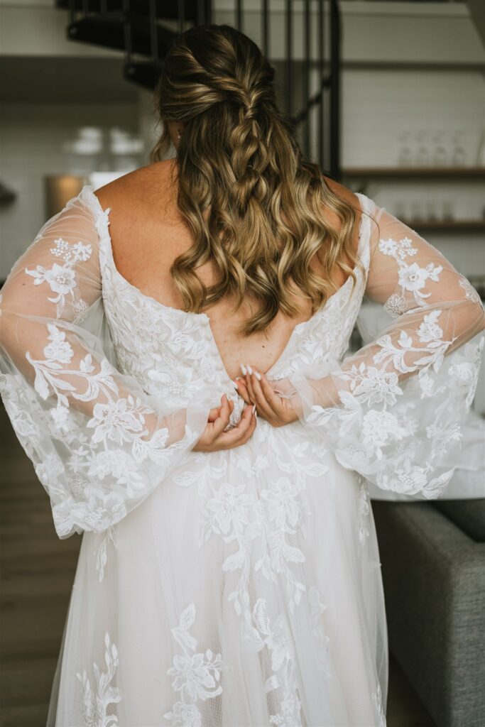 A woman in a lacy white wedding dress stands indoors with her back to the camera, adjusting her gown as she prepares for an intimate lakeside elopement.