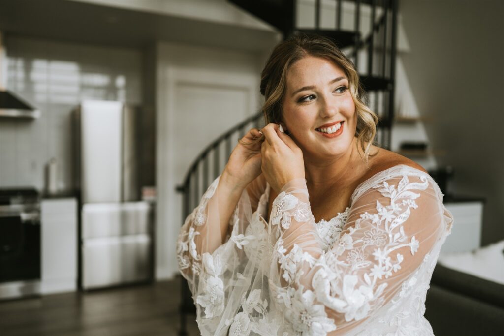 A woman in a white lace dress smiles while putting on an earring, seated in a modern kitchen with a spiral staircase—a serene moment before her lakeside elopement.