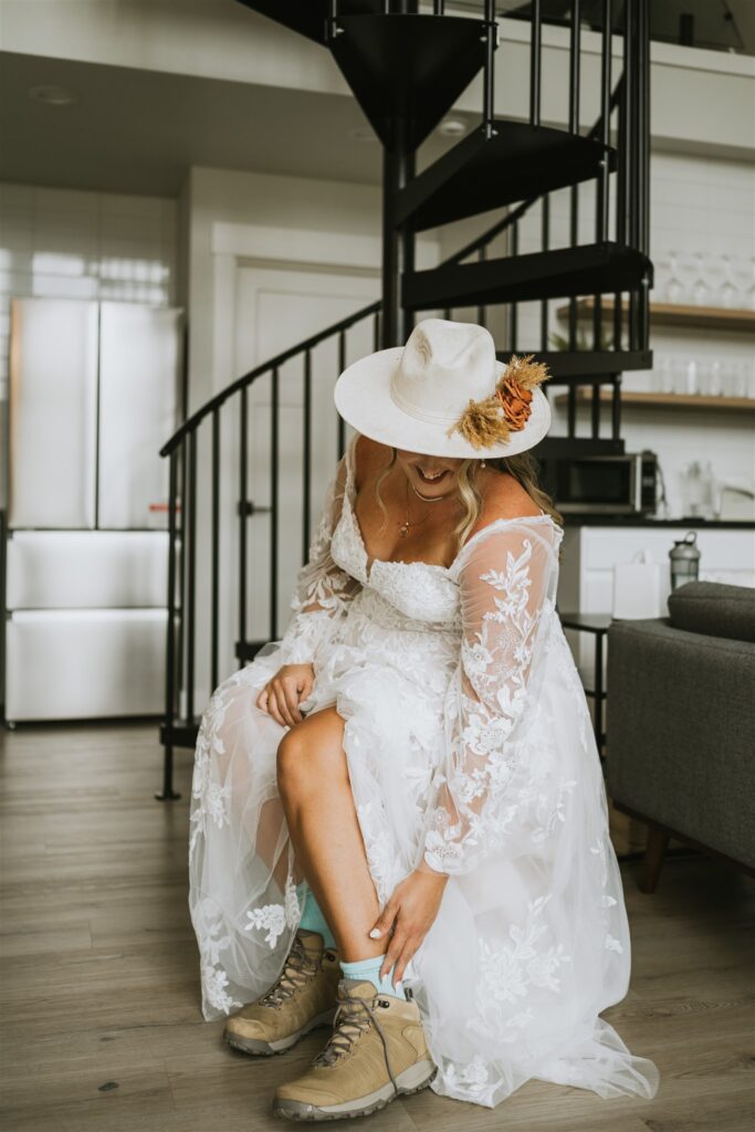 A bride in a lace wedding dress and white hat smiles while putting on tan hiking boots, seated near a spiral staircase in a modern living space—perfectly capturing the adventurous spirit of a lakeside elopement.