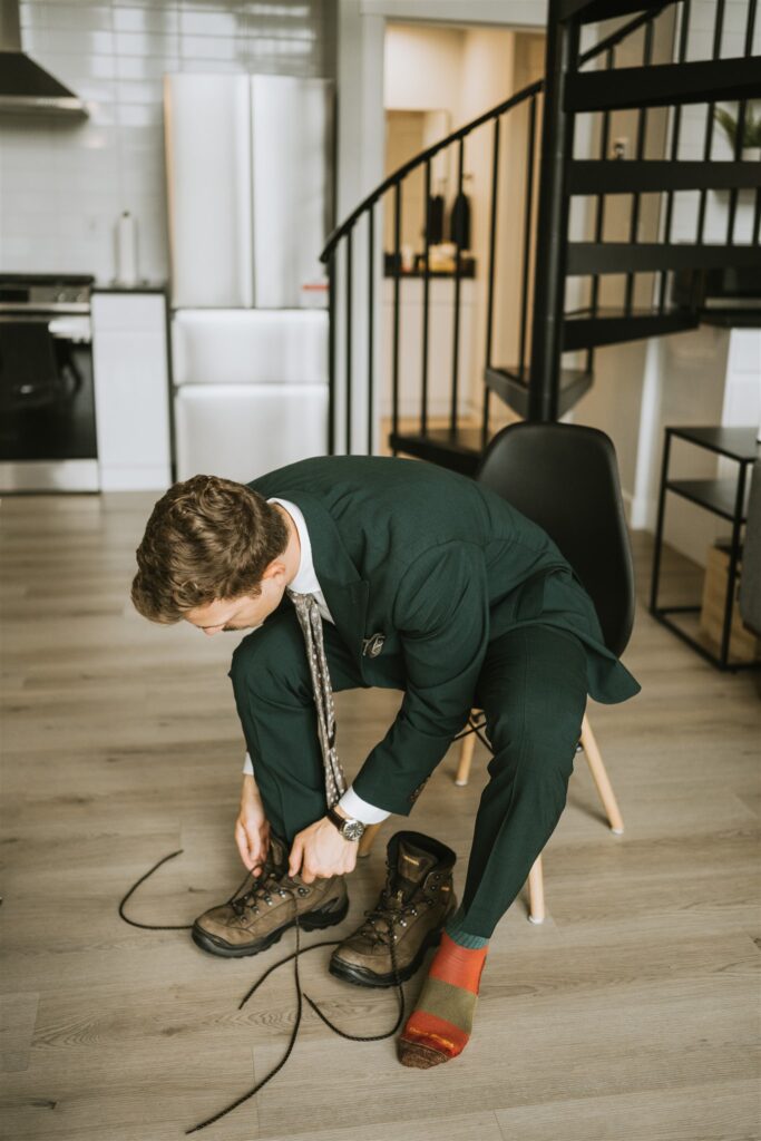 A man in a green suit sits on a chair tying the laces of brown hiking boots, colorful sock visible on his other foot, preparing for a lakeside elopement in a modern kitchen setting.