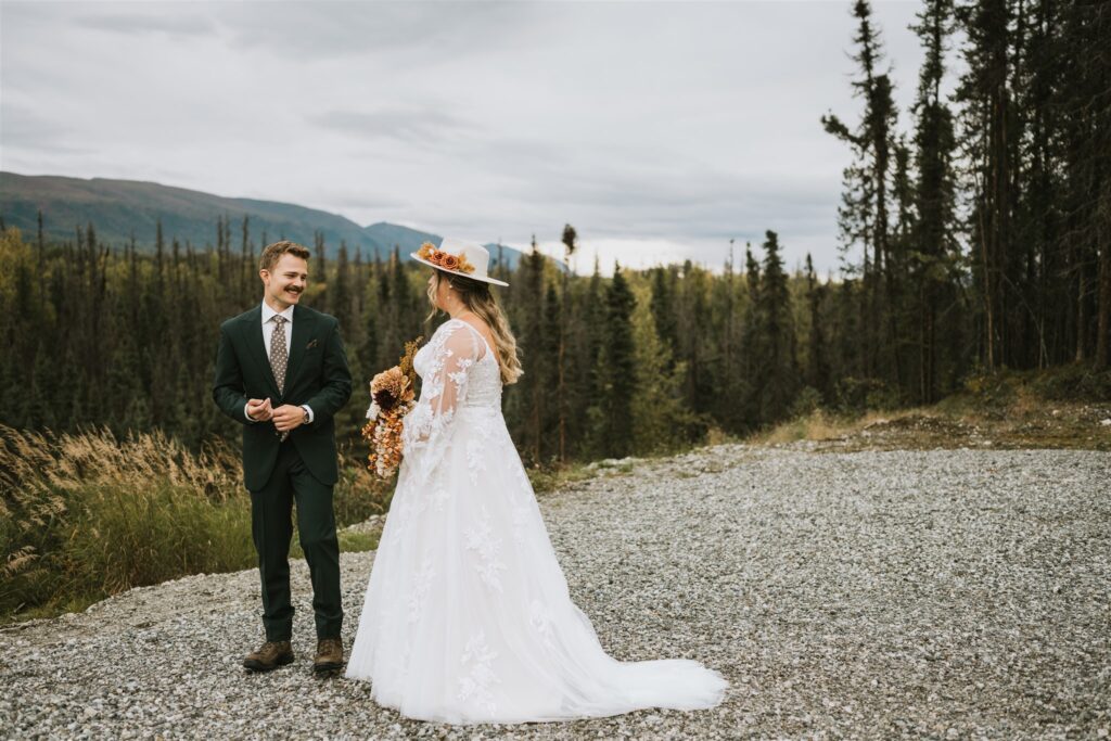 A bride in a white dress and hat holds a bouquet while facing a groom in a dark suit. They stand outdoors on gravel, surrounded by trees and mountains, capturing the intimate charm of a lakeside elopement.