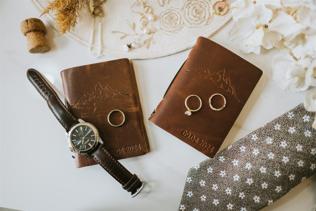 Two brown leather notebooks engraved with mountains and "09.04.2024," a wristwatch, wedding rings, a tie, cork, earrings, and white flowers are artfully arranged on a table—perfect keepsakes for a lakeside elopement.