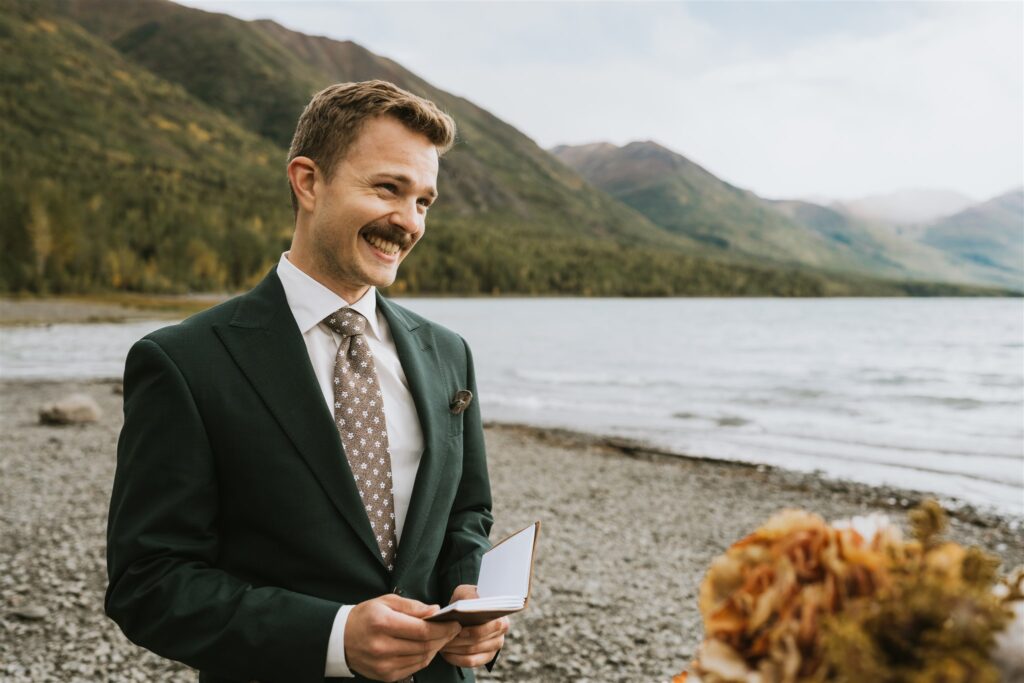 A man in a suit stands by a lake holding an open notebook, smiling, with mountains and trees in the background—capturing the joy of a lakeside elopement.