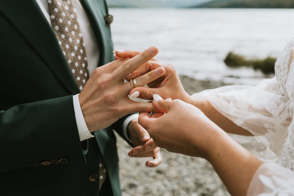 A person in a dark suit places a ring on another’s finger during a romantic lakeside elopement, with the tranquil lake and shore in the background.