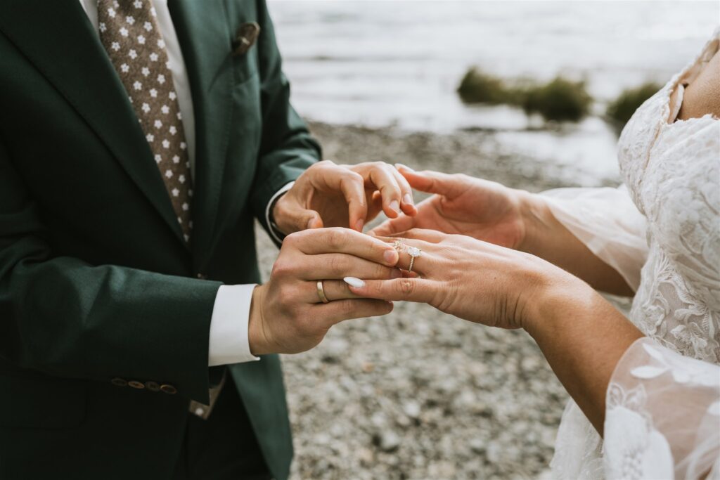A person in a dark green suit places a ring on another's finger during a lakeside elopement by a rocky shoreline.