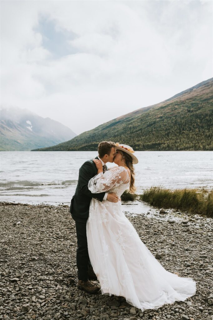 A bride and groom share a kiss on a rocky lakeshore, surrounded by mountains, water, and a cloudy sky—the perfect scene for a romantic lakeside elopement.
