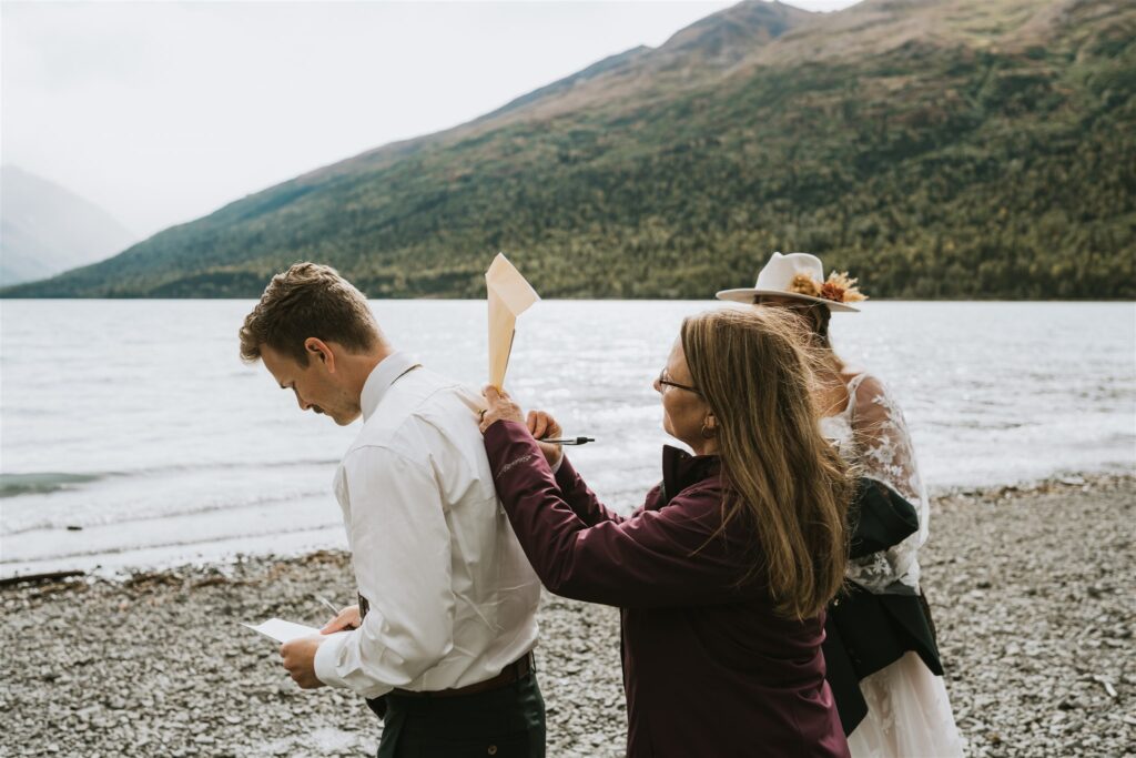 A woman pins a piece of paper to a man's back during a playful lakeside elopement on a rocky shore, while another person in a hat stands nearby.