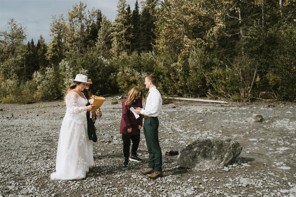 Three people stand on a rocky outdoor area among trees for a lakeside elopement; one in a wedding dress, one in a suit, and one holding papers, appearing to officiate the ceremony.