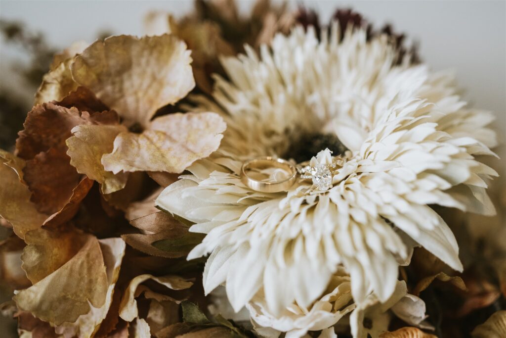 A gold wedding band and a diamond engagement ring rest on a white flower, surrounded by dried brown and beige petals—perfect details for a romantic lakeside elopement.
