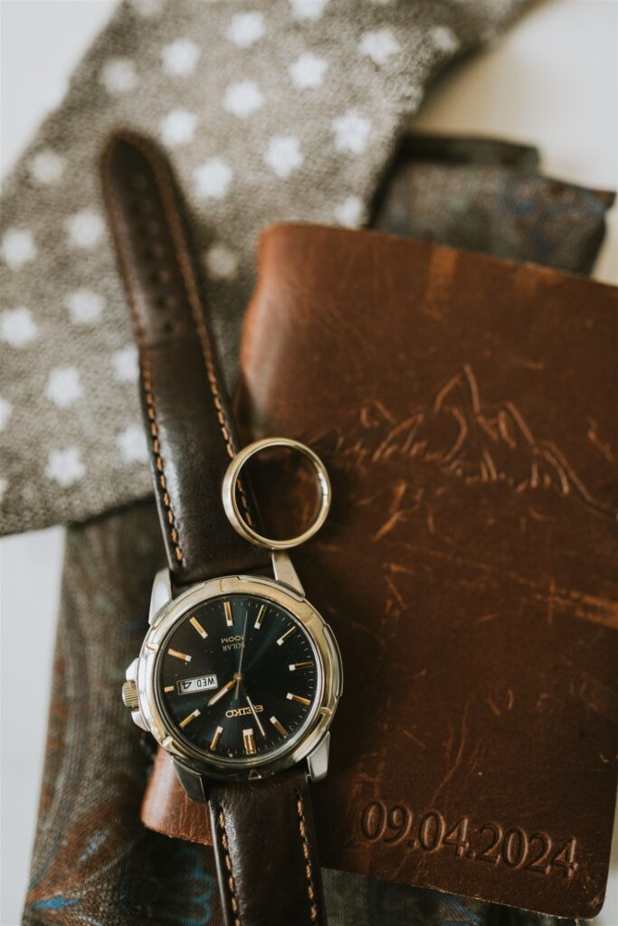 A wristwatch, a gold ring, a brown leather wallet embossed with "09.04.2024," and patterned fabric are arranged together, evoking memories of a lakeside elopement.