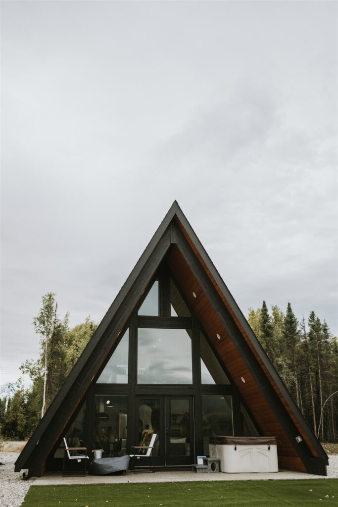 A-frame cabin with large glass windows, patio chairs, and a hot tub—perfect for a lakeside elopement—set against trees under an overcast sky.