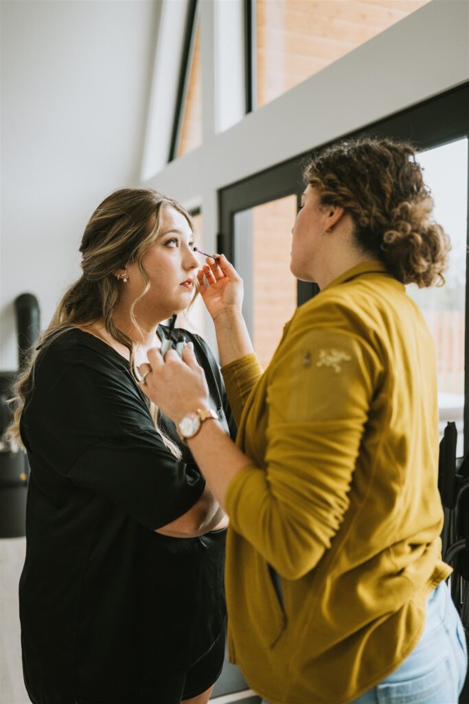 A woman applies makeup to another woman standing by a large window in a well-lit room, preparing for an intimate lakeside elopement.