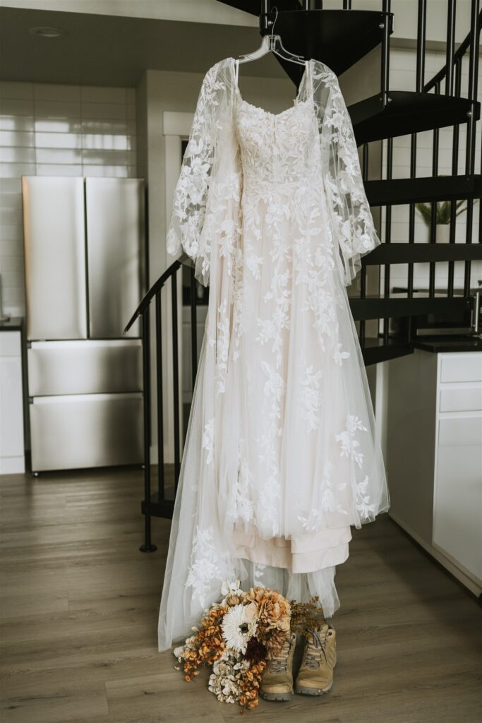 A white lace wedding dress hangs on a hanger near spiral stairs, with a bouquet of flowers and a pair of boots placed on the floor beneath it—perfect for an intimate lakeside elopement.