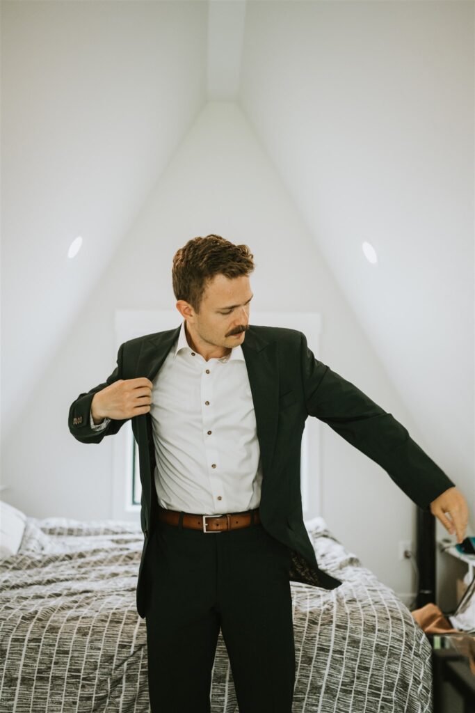 A man with a mustache puts on a dark suit jacket in a bedroom with a white sloped ceiling and neatly made bed, preparing for a lakeside elopement.